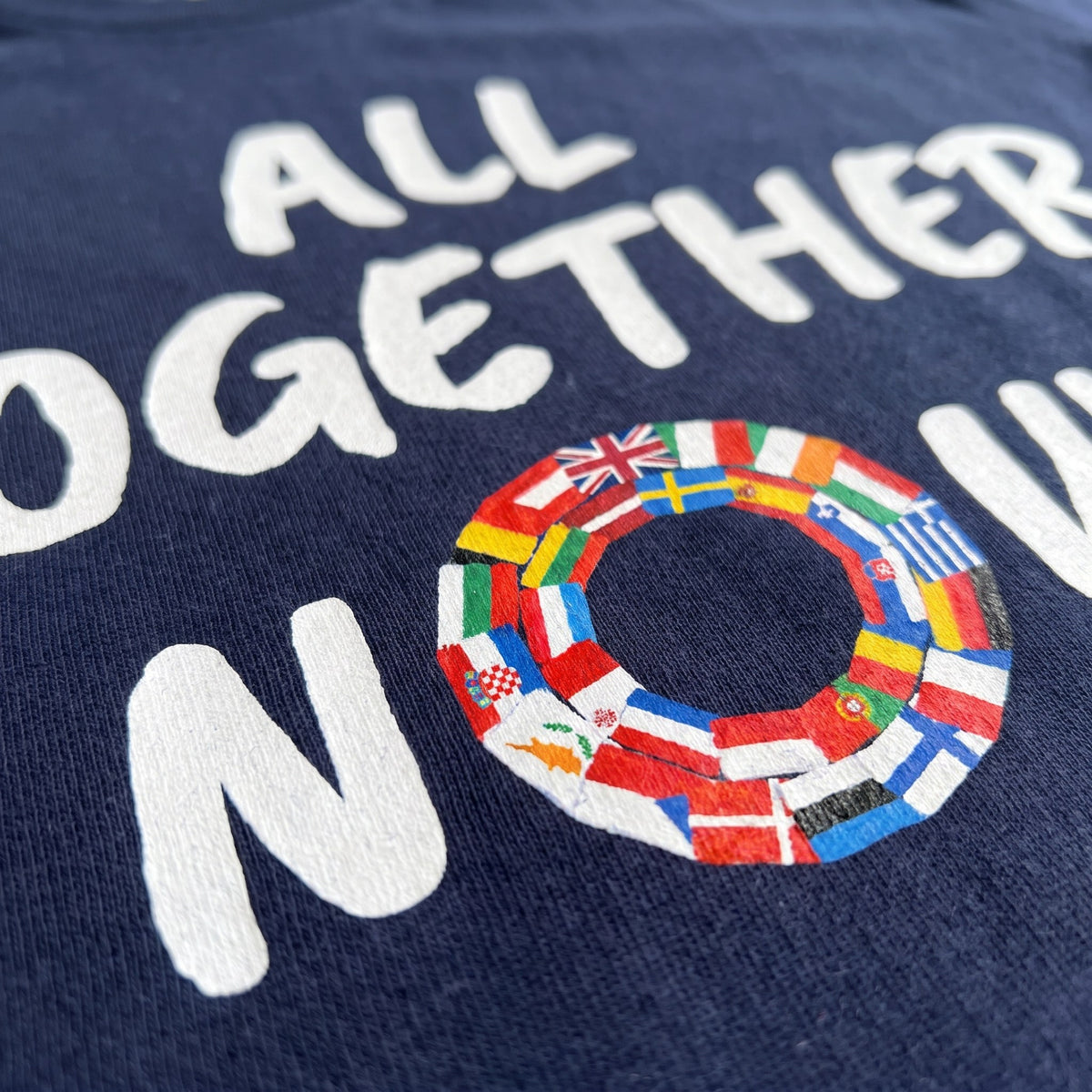 Close-up of the All Together Now print on a navy t-shirt, the ring of European flags forming the O, showing cotton texture and ink detail