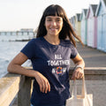 Woman on a seaside promenade wearing the navy All Together Now t-shirt, a ring of 28 hand-painted flags as the O