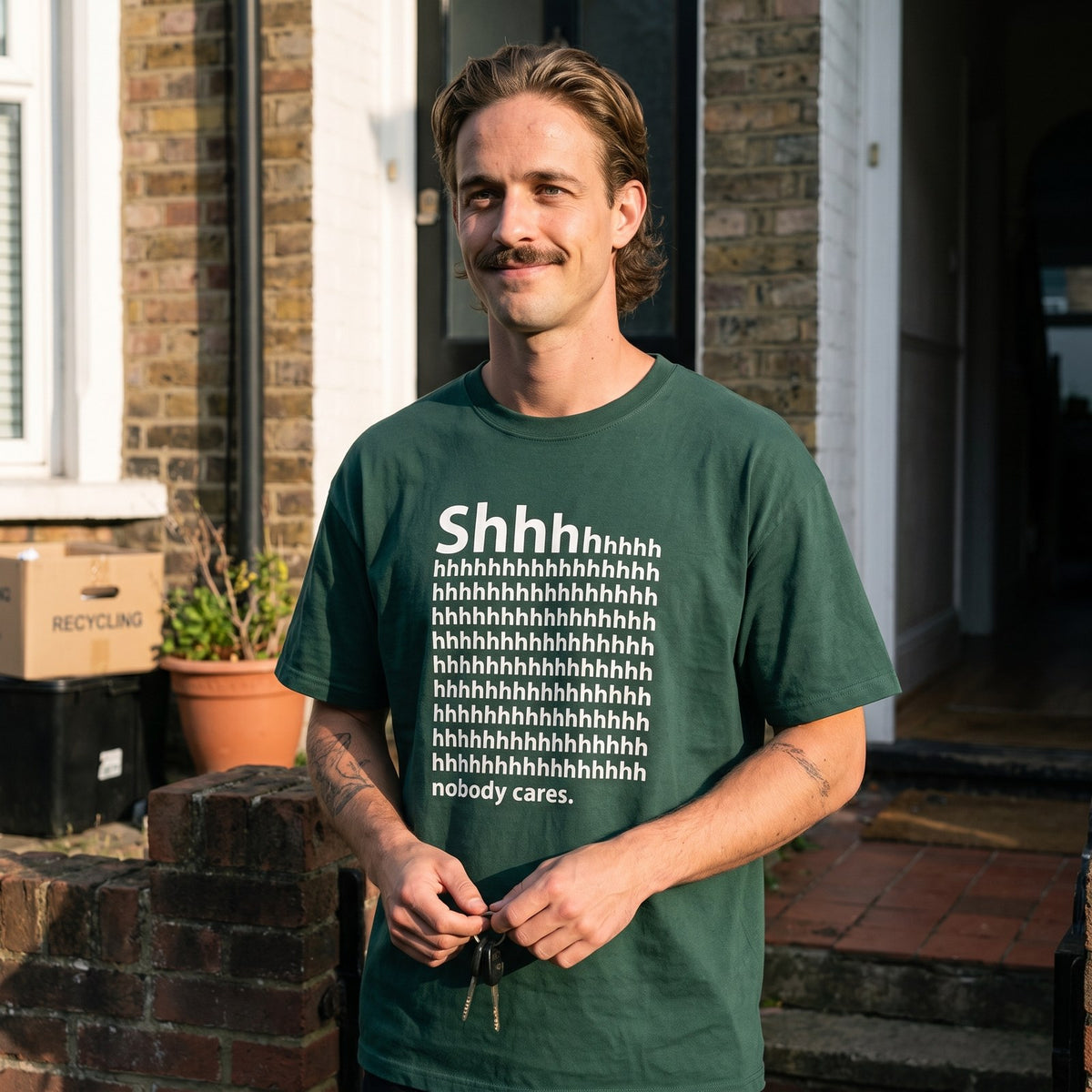Man in a forest green Shhh Nobody Cares t-shirt on a front doorstep