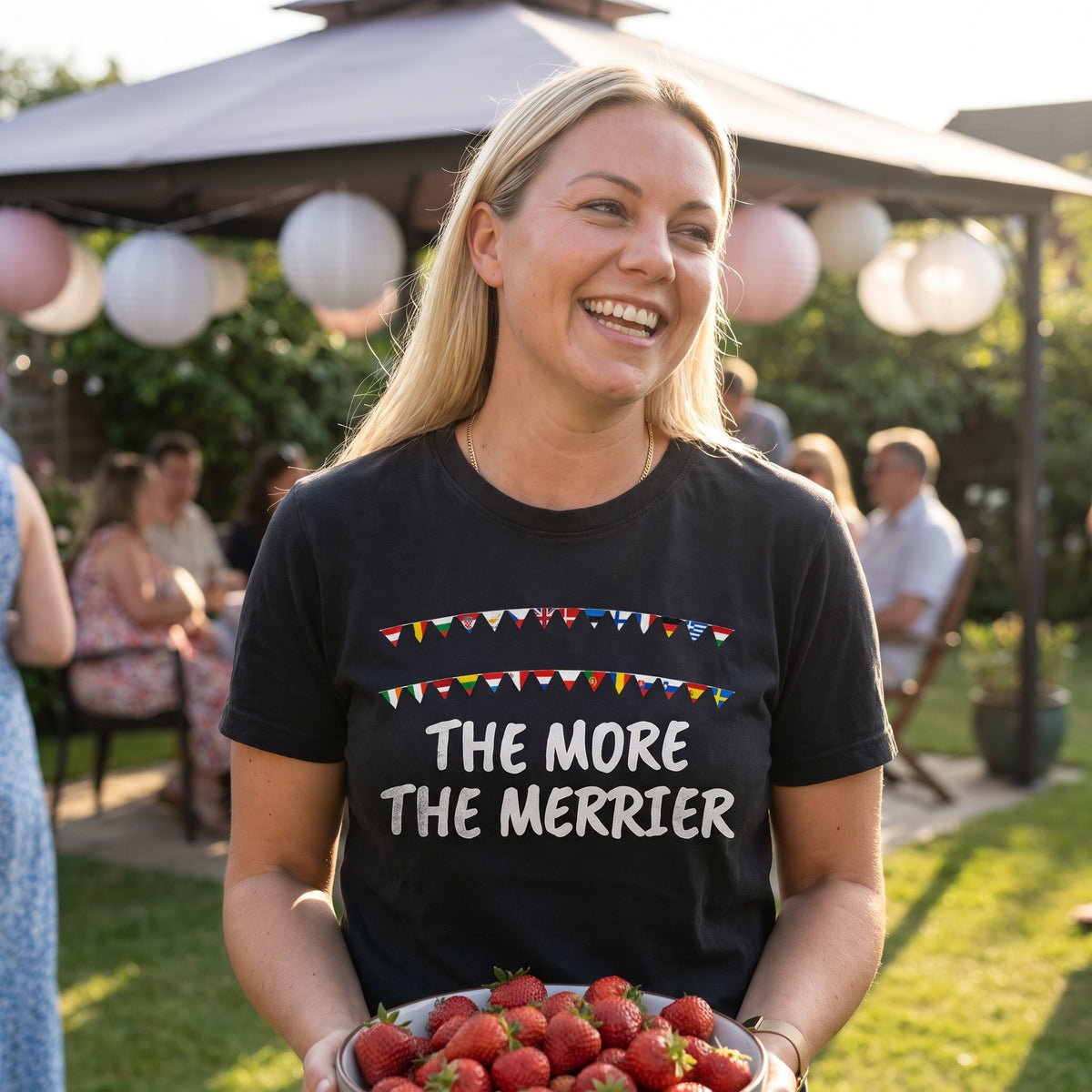 Woman with strawberries at a garden party wearing the black The More the Merrier t-shirt