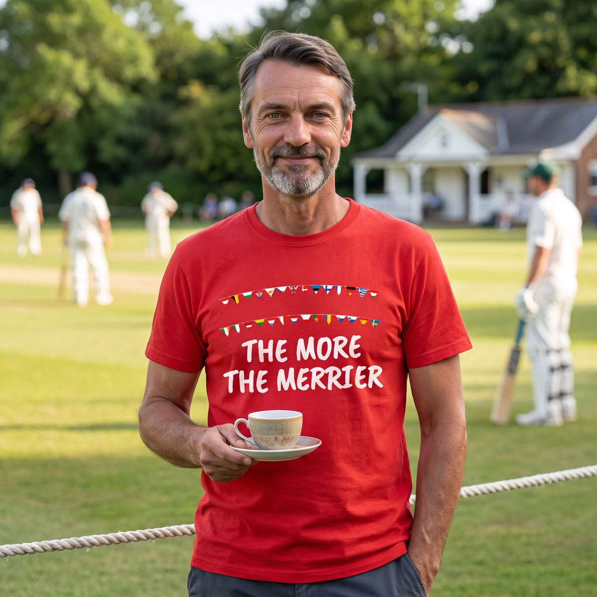 Man with a cup of tea at a village cricket match wearing the red The More the Merrier t-shirt