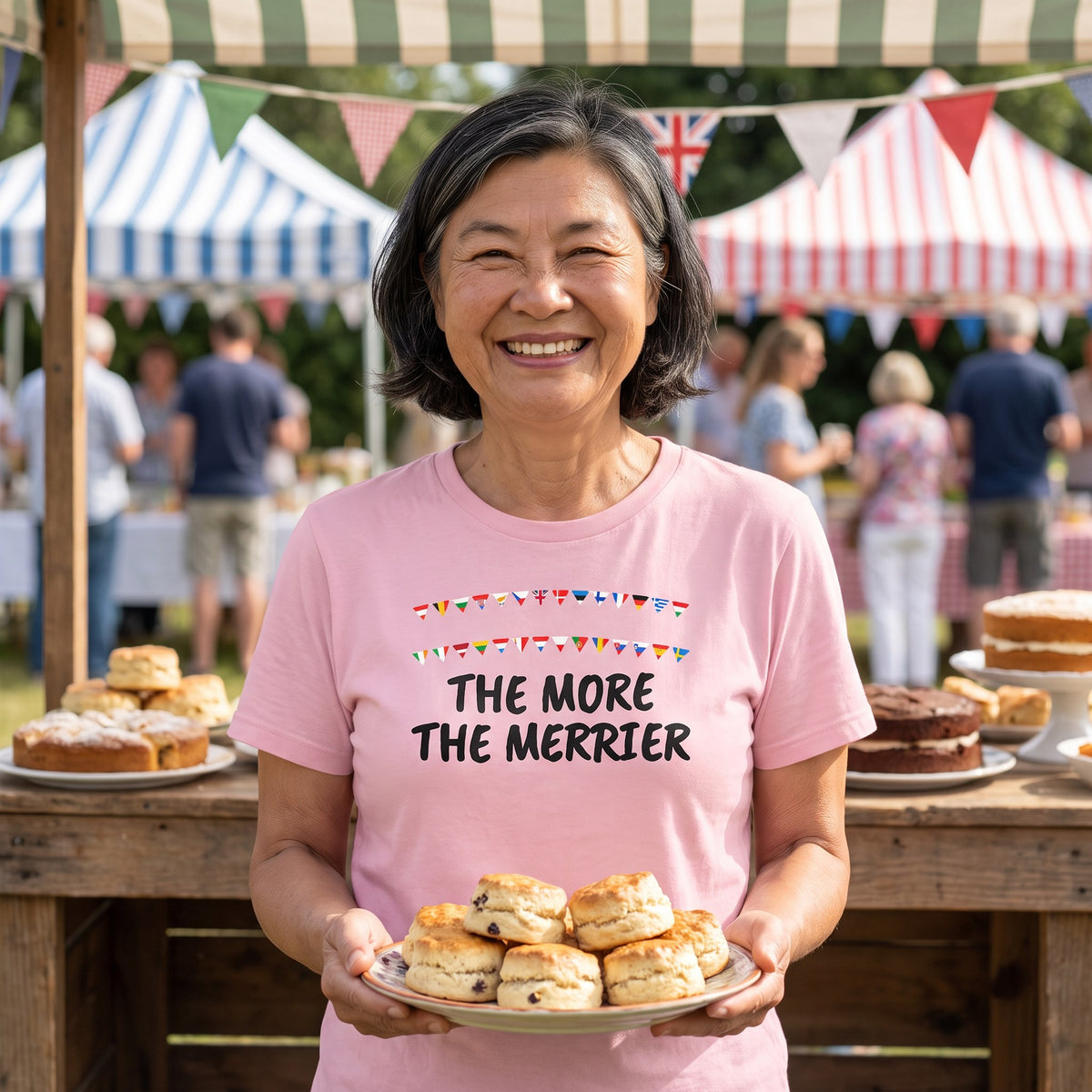 Woman holding scones at a village fete wearing the pink The More the Merrier t-shirt
